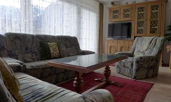 A cozy living room with a sofa and two armchairs. A wooden table sits on a red carpet, and large windows let in plenty of light.