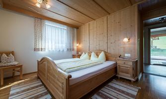 A cozy bedroom with wooden paneling and a large window. The bed is decorated with yellow pillows, and there is a small table with a chair.