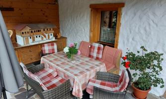 A cozy outdoor area with a table and chairs. On the table, there is a flower vase, and next to a window, there are decorative plants.
