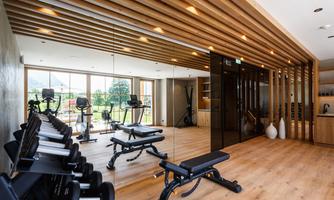 A modern gym with wooden paneling and large windows. There are various workout machines and plenty of daylight.