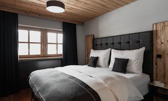 A modern bedroom with a large bed and a wooden ceiling. The walls are painted in neutral colors and large windows provide natural light.