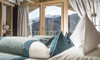 An elegant room with a large window that shows a beautiful mountain landscape. The decor is modern and cozy, with cushions in soft colors.