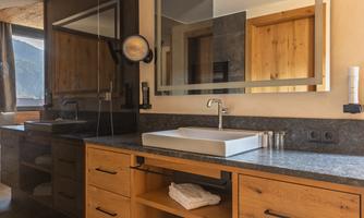 A modern bathroom with wooden details and a large mirror. The sink countertop is made of dark stone and there are towels on a shelf.