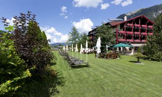 A beautiful hotel building surrounded by a well-maintained lawn. Loungers and sun umbrellas invite you to relax, while the mountains are visible in the background.