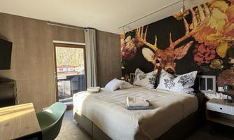 A modern hotel room with a large bed and striking wall decor featuring deer motifs. The room has a window and a table with a chair.
