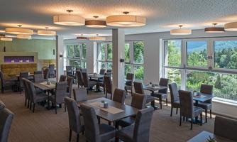 A modern restaurant with bright windows and a beautiful view of greenery. The tables are cozily set, and the decor is stylish and inviting.