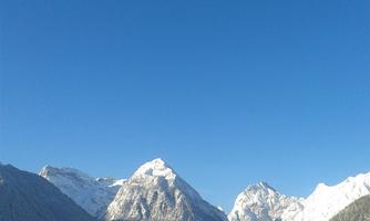 A winter landscape with snow-covered mountains and a clear blue sky. In the foreground, a tranquil lake can be seen.