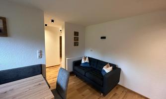 A modern living room with a dark sofa and a wooden chair table. The walls are light and the floor is made of wood, creating a cozy atmosphere.