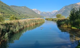 A tranquil river with clear water, surrounded by green meadows and mountains. The sky is blue and radiant.