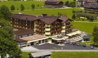 A modern hotel in a picturesque setting with green meadows and mountains in the background. The building features an inviting terrace and already parked cars.