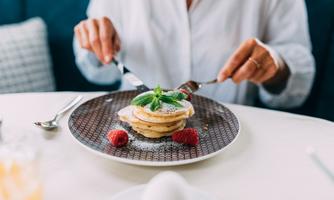 A plate with pancakes, garnished with fresh strawberries and mint. A person is cutting the pancakes with a knife and fork.