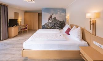 A modern hotel room with two single beds and light-colored furniture. On the wall, there is a picture of a goat on a mountain peak.
