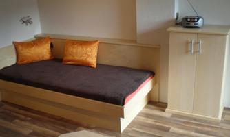 A simple room with a bed and two orange cushions. On the wall, there is a small wardrobe and a radio.