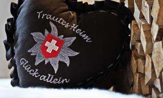 A heart-shaped cushion in black with a Swiss flag and the inscription "Home sweet home, happiness alone." Firewood is visible in the background.