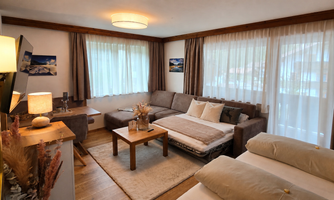 A cozy living room with a beige sofa and a wooden coffee table. Large windows provide plenty of daylight and a warm atmosphere.