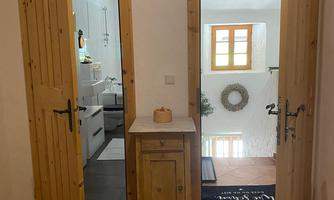 A cozy hallway with open wooden doors. On the left side, a bathroom is visible, while on the right side, a room with a window and a decorative wreath can be seen.