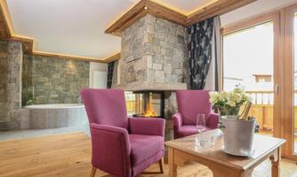 A cozy living room with two purple armchairs and a stone fireplace. A small table with a flower vase and a glass is in the middle.