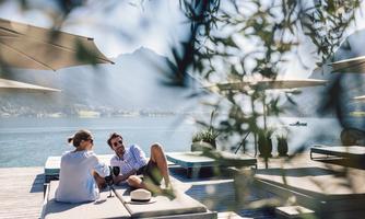 A relaxing moment by the water with two people on loungers. In the background, there are sun umbrellas and a beautiful view of the lake and the mountains.