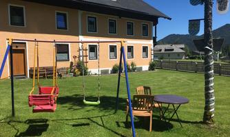 A playground with swings is located in the greenery in front of a yellow house. In the background, there are mountains and a clear blue sky.