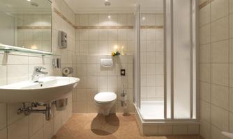 A modern bathroom with white tiles and a walk-in shower. There is a sink and a toilet as well as a wall mirror and lighting.