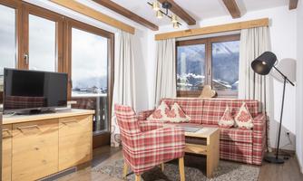 A cozy living room with a red checkered sofa and a coffee table. Large windows let in daylight and offer a view of the mountains.