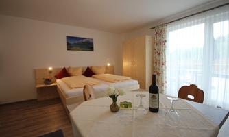A cozy room with a double bed and a dining area. On the table, there is a bottle of wine and a vase with flowers.