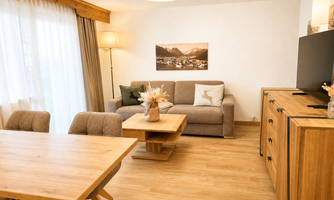 A modern living room with wood and neutral tones. There is a cozy sofa, a wooden table, and large windows that let in plenty of light.