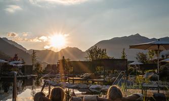 A relaxed scene by the pool with two loungers and a stunning view of the mountains. The sun sets behind the mountains, creating a warm, inviting atmosphere.