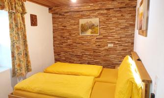 A cozy bedroom with two yellow beds and rustic wooden decor. The walls are clad with stone slabs, and there are windows with floral curtains.