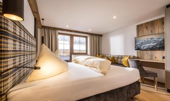 A modern bedroom with a large bed and wooden elements.  
Lots of light comes through the windows, and there is a television as well as comfortable seating options.