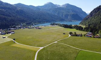 A vast meadow landscape with mountains in the background. A tranquil lake and small paths are also visible.