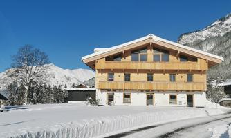 A modern wooden house in a snowy landscape. In the background, mountains and blue sky are visible.