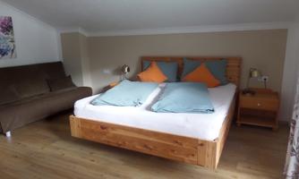 A cozy bedroom with a wooden double bed and colorful cushions. Next to the bed is a light brown nightstand and a sofa in the corner.