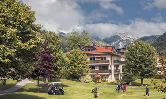 A picturesque golf course surrounded by trees and mountains. In the foreground, golfers are playing on a green meadow.