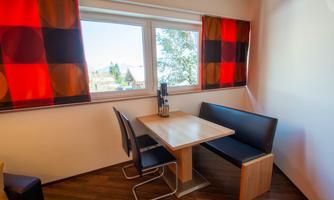 A cozy dining area with a table and a bench. The windows are adorned with colorful curtains and offer a view of the outside world.