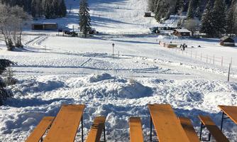A snow-covered landscape with a ski slope in the background. In the foreground, rustic wooden tables and benches stand.