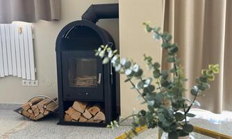 A modern fireplace stands in a bright room. Next to the fireplace is a stack of firewood and a small table with a plant.