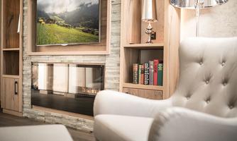 A modern living room with a comfortable white armchair and a television on the wall. In the background, a fireplace and bookshelves are visible.