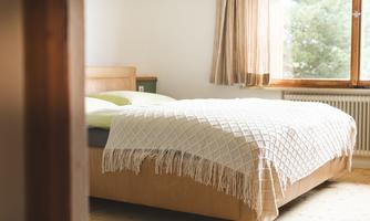 A cozy bedroom with a large bed and a white bedspread. The windows let in plenty of natural light.