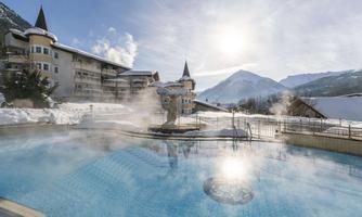 A heated outdoor pool in a wintry landscape with steaming water. In the background, snow-covered mountains and a radiant sky can be seen.