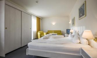 A modern hotel room with a cozy double bed and a seating area. The walls are light and there are large windows with curtains.