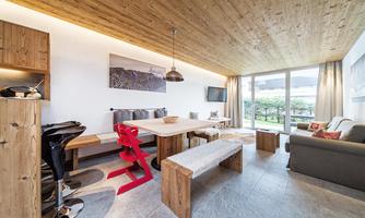 A modern living room with a wooden ceiling, a dining area, and a cozy seating nook. Large windows let in plenty of daylight and offer a view of the outdoors.