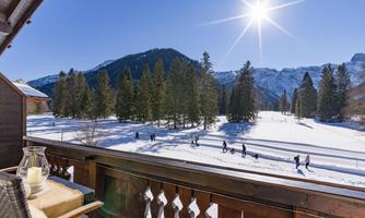A snowy landscape with tall fir trees and majestic mountains in the background. The sun shines bright in the sky and people walk along a path.
