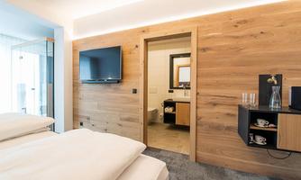 A modern hotel room with wooden paneling and a cozy bed. In the background, the bathroom and a television are visible.