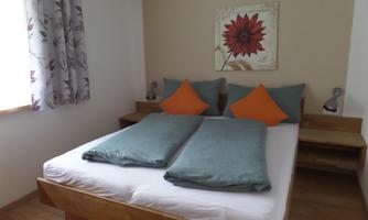 A cozy bedroom with a double bed and colorful cushions. A flower picture hangs on the wall, and light falls through the window.