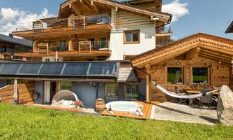 A cozy wooden house with several balconies and a beautiful environment. In the foreground, there is an outdoor seating area with a bathtub.