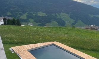 A modern wooden pool with a view of a green mountain landscape. The sky is partly cloudy and nature looks idyllic.