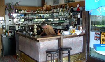 A cozy bar with a bartender working at the counter. In the background, numerous bottles and drinks are visible.