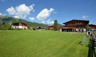 A green meadow surrounded by modern buildings in a picturesque mountain landscape. The sky is blue with a few white clouds.