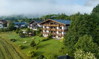 A cozy hotel in a green landscape, surrounded by trees and flowers. In the background, there are gentle hills and a clear sky with some mist.